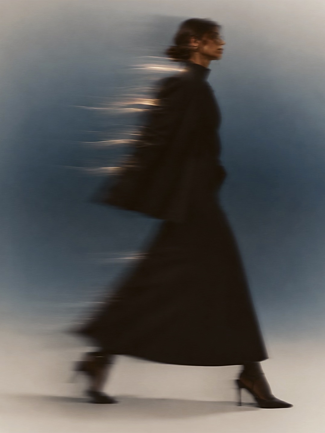 A figure in a long dark coat and heels, captured mid-stride against a blue-grey backdrop.